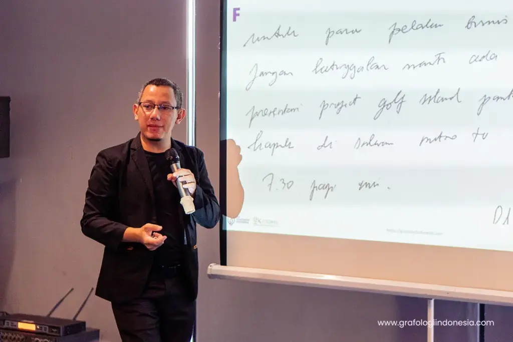 Kang Aviv memberikan presentasi di depan layar yang menampilkan tulisan tangan dengan kata-kata dalam Bahasa Indonesia.